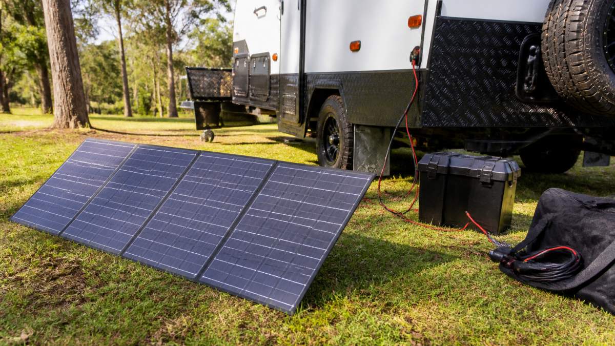 A portable solar panel set up on grass beside a caravan at a bush campsite, with cables running to the van and a battery box visible nearby in warm afternoon light
