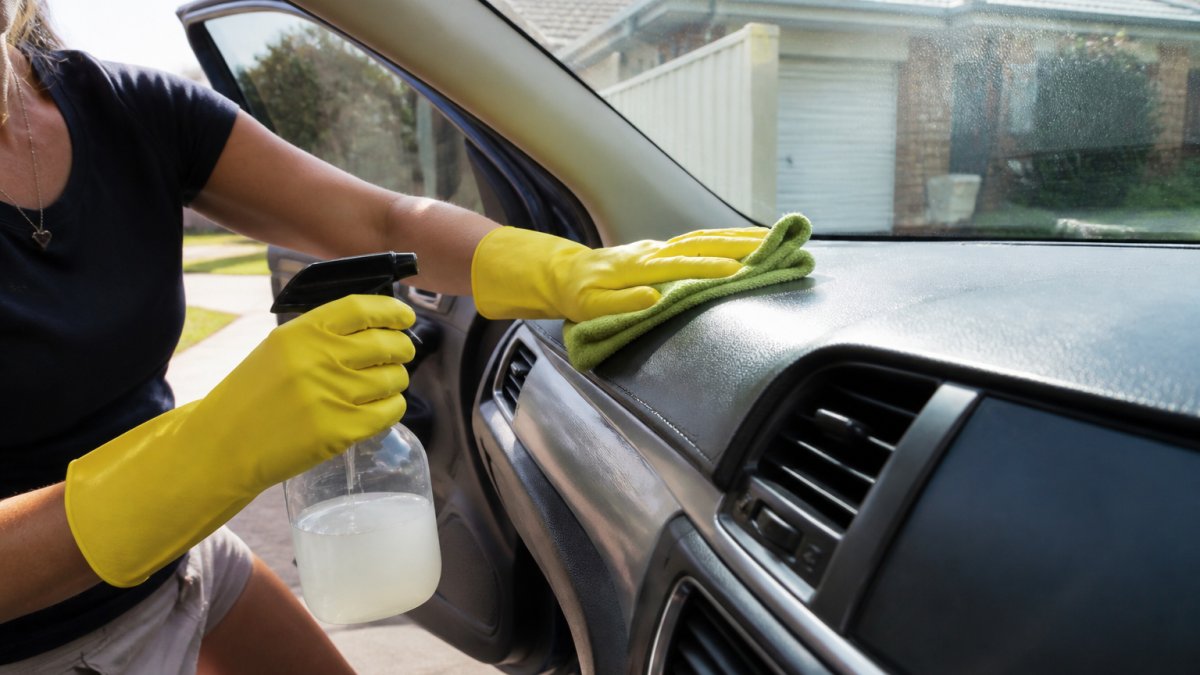 A person wearing rubber gloves wiping a car dashboard with a microfiber cloth and a spray bottle of homemade cleaning solution, car door open in a sunny driveway